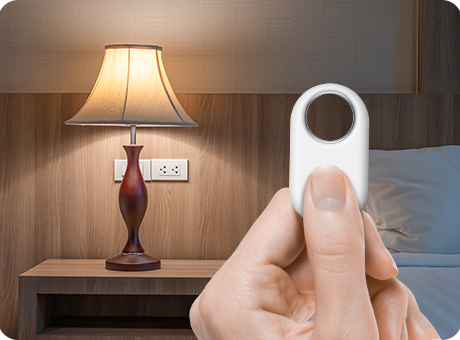 Samsung Galaxy SmartTag2-Tracker in einer Hand. Die Person drückt den Knopf auf dem Gerät. Im Hintergrund ein modern eingerichtetes Schlafzimmer mit eingeschalteter Nachttischlampe.