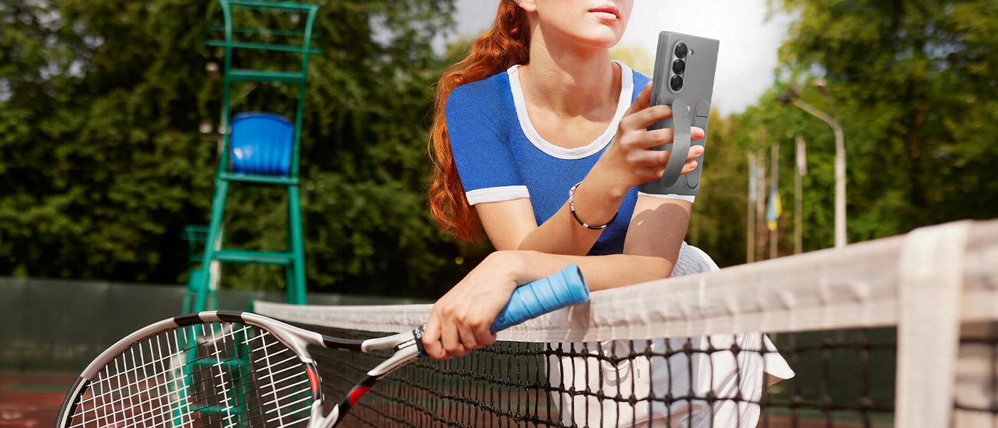 Junge Frau auf einem Tennisplatz lehnt an einem Netz und hält bequem Galaxy Z Fold6. Das Telefon befindet sich in einer grauen Silicone Case, und die Frau hält das Telefon mit einem Halter, der durch ihre Finger geführt wird.